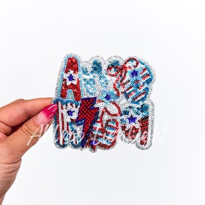 May include: A sparkly red, white, and blue iron-on patch with the word "USA" in a stylized font. The patch is decorated with stars, stripes, and other patriotic imagery.