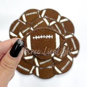 May include: A brown and white felt football patch with stitching details. The patch is held in a hand with black nail polish.