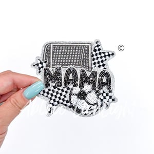 May include: A black and white iron-on patch with the word "MAMA" in large, sparkly letters. The patch also features a soccer ball, a goal, and a checkered star.