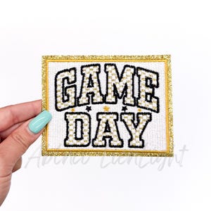 May include: A white embroidered patch with gold glitter trim. The patch says "GAME DAY" in black and gold lettering.