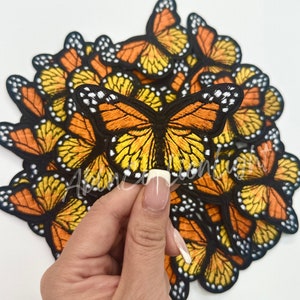 May include: A pile of embroidered butterfly patches in orange, yellow, and black. The patches are arranged in a semi-circle with one patch being held in a hand.