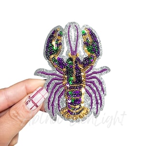 May include: A sequined lobster patch in purple, green, and gold, with a silver glitter border. The lobster design is held by a hand against a white background. The patch is likely for clothing or accessories.