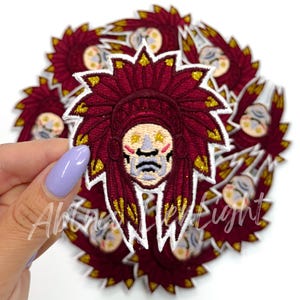 May include: A stack of red and gold embroidered iron-on patches featuring a Native American headdress with a face. The headdress has white feathers and gold glitter accents.