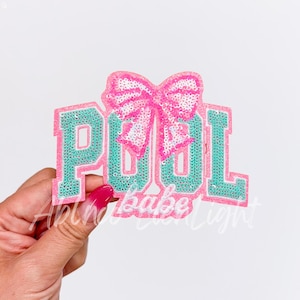 May include: A sequined patch with the word "POOL" in teal letters outlined in pink, topped with a pink sequined bow. The patch has a white background and is held by a hand with a pink nail.