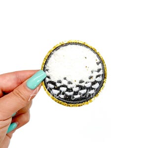 May include: A white and black golf ball patch with a gold glitter border.