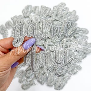 May include: A pile of silver glitter iron-on patches that say "game day" in a cursive font.