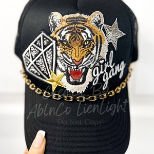 Peut inclure: Casquette de camionneur noire avec un visage de tigre, un diamant et une &eacute;toile brod&eacute;s sur le devant. La casquette a &eacute;galement une bordure en cha&icirc;ne dor&eacute;e et le texte "girl gang" brod&eacute; sur le c&ocirc;t&eacute;.