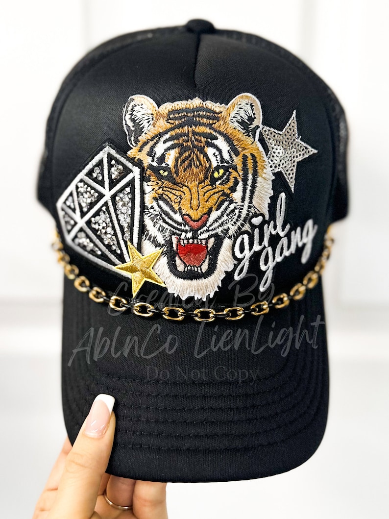 Peut inclure: Casquette de camionneur noire avec un visage de tigre, un diamant et une &eacute;toile brod&eacute;s sur le devant. La casquette a &eacute;galement une bordure en cha&icirc;ne dor&eacute;e et le texte "girl gang" brod&eacute; sur le c&ocirc;t&eacute;.