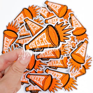 May include: A pile of orange and white iron-on patches featuring a megaphone with the word "CHEER" on it. The megaphone has a white outline and a white starburst behind it.
