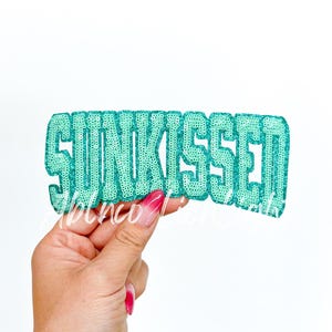May include: A teal blue iron-on patch with the word "SUNKISSED" in large, glittery letters.
