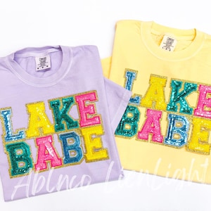 May include: Two t-shirts with the text "LAKE BABE" in colorful sequins. The shirts are in lavender and yellow.
