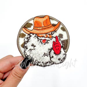 May include: Embroidered iron-on patch of a cartoon cowboy Santa Claus wearing a brown cowboy hat with an orange band, a white beard, and a red glove. The patch is on a brown and white cowhide background.