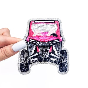 May include: A pink and silver sequined patch in the shape of an off-road vehicle. The patch has a glittery silver border and the text "Rhino Lifestyles" in a cursive font.