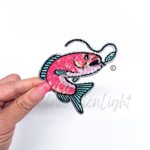 May include: A pink and blue sequined fish with a silver lure in its mouth. The fish is embroidered with sequins and has a silver glitter finish.