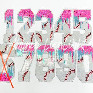 May include: A collection of baseball-themed number patches, each featuring a silver sequined base. The numbers are adorned with pink glitter, blue drips, and red baseball stitching. The numbers range from 1 to 0, with the number 6 crossed out.