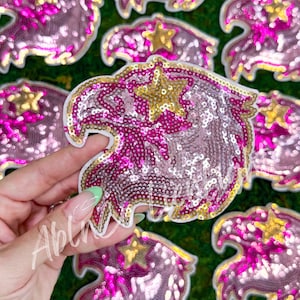 May include: Pink and gold sequined iron-on patches in the shape of an eagle with a star on its chest.