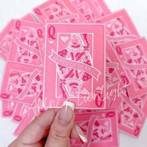 Puede incluir: Un parche bordado rosa de la carta de juego de la Reina de Corazones. El parche presenta un contorno blanco de la Reina sosteniendo una espada y un coraz&oacute;n. El texto "Queen of Hearts" est&aacute; escrito en una pancarta blanca.
