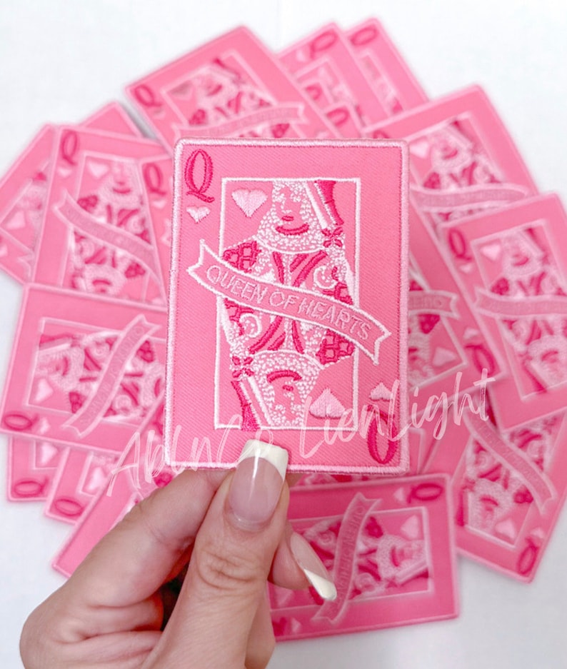 Puede incluir: Un parche bordado rosa de la carta de juego de la Reina de Corazones. El parche presenta un contorno blanco de la Reina sosteniendo una espada y un coraz&oacute;n. El texto "Queen of Hearts" est&aacute; escrito en una pancarta blanca.
