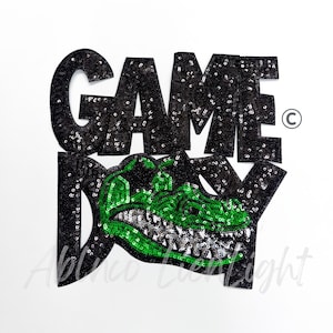 May include: Black sequined "GAME DAY" applique with a green alligator design. The alligator has silver teeth and is outlined in green sequins. The text is in a bold, blocky font.
