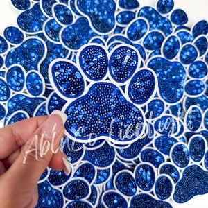May include: A pile of blue and white paw print patches with sequins. The patches are made of fabric and have a white outline. The sequins are blue and create a sparkly effect.