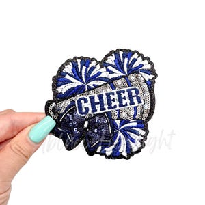 May include: Blue and white cheerleading pom poms with a silver sequined megaphone and the word "CHEER" in black letters.