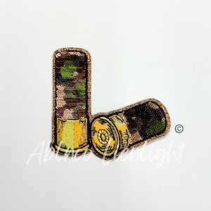 May include: A pair of gold and green camouflage shotgun shells. The shells are made of fabric and have a glittery finish.