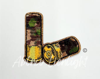 May include: A pair of gold and green camouflage shotgun shells. The shells are made of fabric and have a glittery finish.