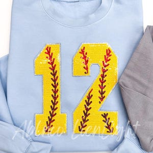 May include: Light blue sweatshirt with a large sequined number 12 in yellow with red stitching. The number is designed to look like a softball.