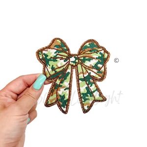 May include: A decorative bow with a camouflage pattern in shades of green, brown, and white. The bow has a brown outline with a glittery texture. The bow is held by a hand with a light blue nail polish.