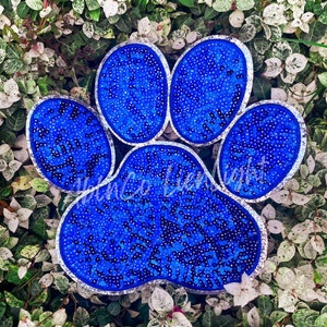 May include: A large blue paw print made of sequins with a silver outline. The paw print is on a background of green leaves.