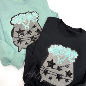 May include: Two sweatshirts with a cauldron design. One sweatshirt is teal green and the other is black. The cauldron is decorated with stars and flowers.