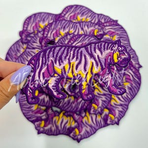 May include: A set of purple and yellow embroidered iron-on patches featuring a tiger design. The patches are in the shape of a circle and have a textured, fuzzy surface.