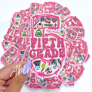 May include: Pink felt patch with the number 5 and the words "Fifth Grade" in a glittery pink font. The patch is decorated with school-themed illustrations, including a pencil, a backpack, and a lightning bolt.