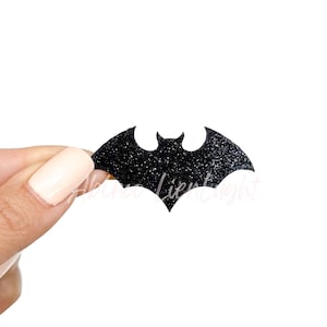 May include: A black glitter bat-shaped embellishment. The bat has a classic silhouette with pointed ears and spread wings. The surface is covered in sparkling glitter, catching the light. This decorative element is ideal for Halloween or themed crafts.