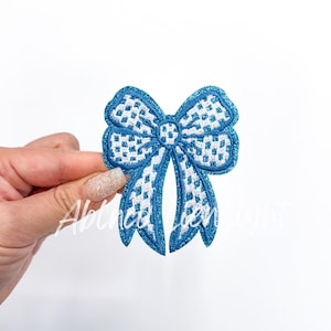May include: A blue and white bow applique with a glittery border. The bow features a detailed, openwork design. The text "Ablinca den light" is visible on the lower left side of the bow.