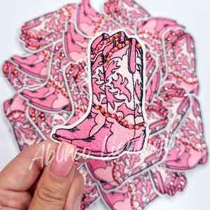 May include: Pink embroidered cowboy boot patch with rhinestones. The patch is surrounded by other pink boot patches.