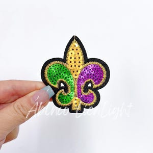 May include: A gold, green, and purple sequined fleur-de-lis patch. The patch is on a black felt background.