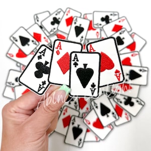 May include: A hand holds three playing cards, each an Ace, with a suit of clubs, diamonds, and spades. The cards are white with black and red embroidery. The cards are surrounded by a pile of other playing cards, all with different suits and values.