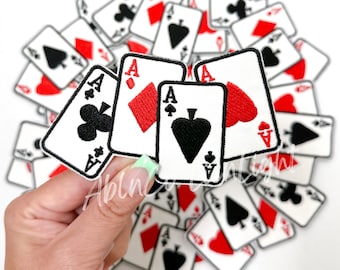 Puede incluir: Una mano sostiene tres cartas de juego, cada una un As, con un palo de trébol, diamantes y picas. Las cartas son blancas con bordados negros y rojos. Las cartas están rodeadas por una pila de otras cartas de juego, todas con diferentes palos y valores.