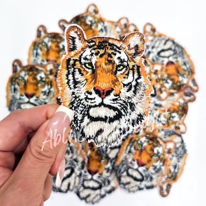 May include: A close-up of a tiger face embroidered patch. The patch is orange, black, and white with a detailed design. The tiger has a fierce expression with sharp teeth and a long, flowing mane.