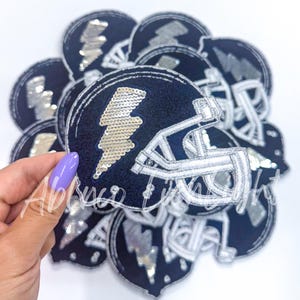 May include: A pile of navy blue and silver iron-on patches featuring a football helmet with a lightning bolt design. The patches are made of felt and sequins.