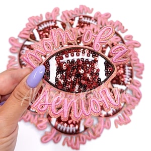 May include: A pink and gold glitter iron-on patch with a football design and the text "Mom of a Senior".