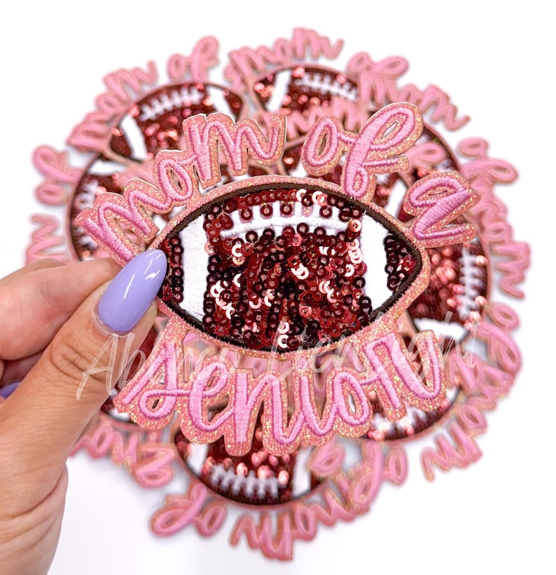 May include: A pink and gold glitter iron-on patch with a football design and the text "Mom of a Senior".
