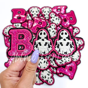 May include: A set of pink and white iron-on patches with the word "BOO" and ghost designs. The patches are made of sequins and fabric.