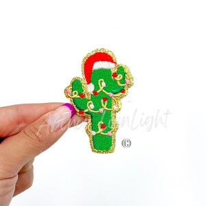 May include: A green cactus wearing a red Santa hat with gold glitter trim and white pom pom. The cactus is decorated with pink and white Christmas lights.