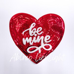 May include: Red heart-shaped sequin patch with white embroidered text that reads "be mine".