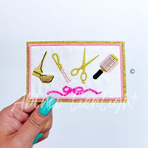 May include: A white embroidered patch with gold glitter trim. The patch features a pink ribbon, a pair of gold scissors, a gold hair clip, a gold hair brush, and a gold hair bowl.