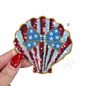 May include: A sequined seashell-shaped patch with a patriotic design. The patch features red and white stripes, a blue bow with white stars, and a gold border. The design is reminiscent of the American flag.
