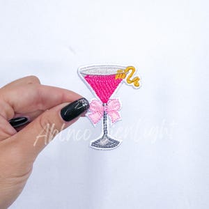 May include: Pink embroidered martini glass with a gold swirl and a pink bow. The glass is filled with a pink liquid.