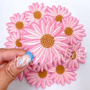 May include: A pile of pink daisy flower iron-on patches with a white center and a brown center. The patches are about 2 inches in diameter.